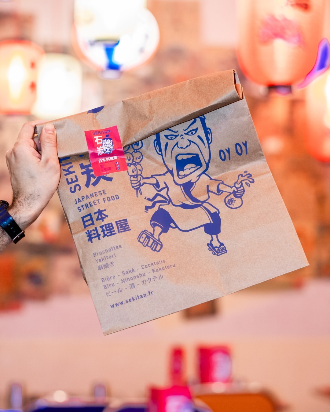 Hand holding a Sekitan Japanese street food block bottom paper bag with bold blue artwork, set against a colorful backdrop of lanterns